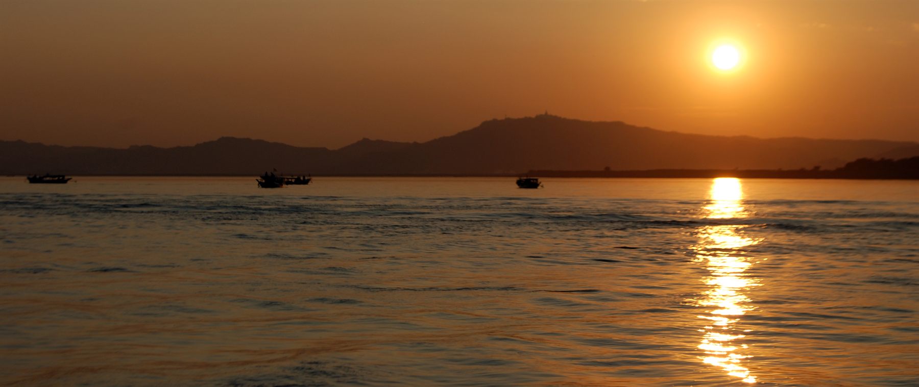 Sun setting over a wide river with small boats silhouetted on the golden water